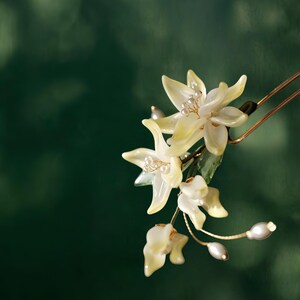 May include: A delicate floral hair accessory featuring creamy yellow flower petals and small pearl accents. The flowers are attached to a gold-toned hairpin, creating an elegant and natural look. The background is a dark green.