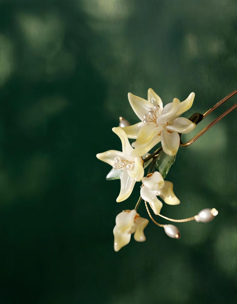 May include: A delicate floral hair accessory featuring creamy yellow flower petals and small pearl accents. The flowers are attached to a gold-toned hairpin, creating an elegant and natural look. The background is a dark green.