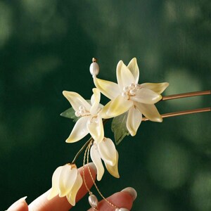 May include: A delicate hair accessory featuring white and yellow flower-shaped ornaments with pearl accents. The floral design includes a gold-colored hairpin and dangling pearl strands, creating an elegant and refined aesthetic. The accessory is held against a dark green background.
