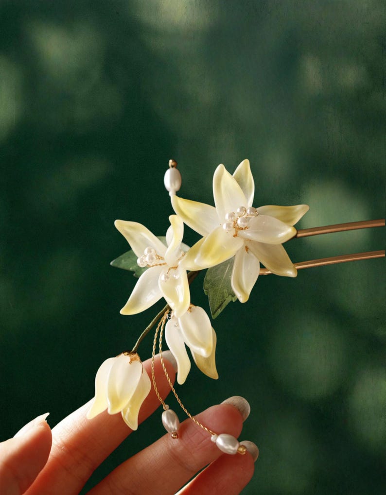 May include: A delicate hair accessory featuring white and yellow flower-shaped ornaments with pearl accents. The floral design includes a gold-colored hairpin and dangling pearl strands, creating an elegant and refined aesthetic. The accessory is held against a dark green background.