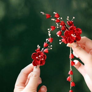 Op de afbeelding: Twee rode bloemen haaraccessoires. E&eacute;n is een clip met een cluster van rode bloemen en witte accenten. De andere is een groter bloemstuk met een gouden ketting en bungelende rode bloemknoppen. Beide tegen een vage groene achtergrond.