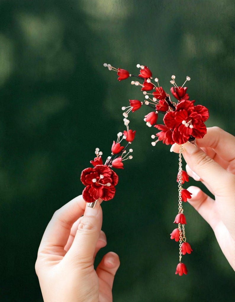 Op de afbeelding: Twee rode bloemen haaraccessoires. E&eacute;n is een clip met een cluster van rode bloemen en witte accenten. De andere is een groter bloemstuk met een gouden ketting en bungelende rode bloemknoppen. Beide tegen een vage groene achtergrond.