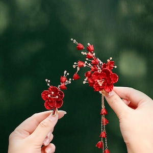 Op de afbeelding: Twee rode bloemen haaraccessoires. E&eacute;n is een enkele bloem met een gouden clip. De andere heeft meerdere bloemen, een gouden ketting en bungelende rode belvormige accenten. Beide worden tegen een vage groene achtergrond gehouden.
