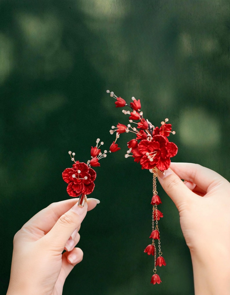Op de afbeelding: Twee rode bloemen haaraccessoires. E&eacute;n is een enkele bloem met een gouden clip. De andere heeft meerdere bloemen, een gouden ketting en bungelende rode belvormige accenten. Beide worden tegen een vage groene achtergrond gehouden.