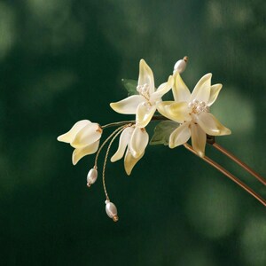 May include: A delicate hair accessory featuring stylized, pale yellow flowers with pearl accents. The floral design includes small green leaves and dangling pearl-like beads, all mounted on a gold-toned metal pin. The background is a blurred, dark green.