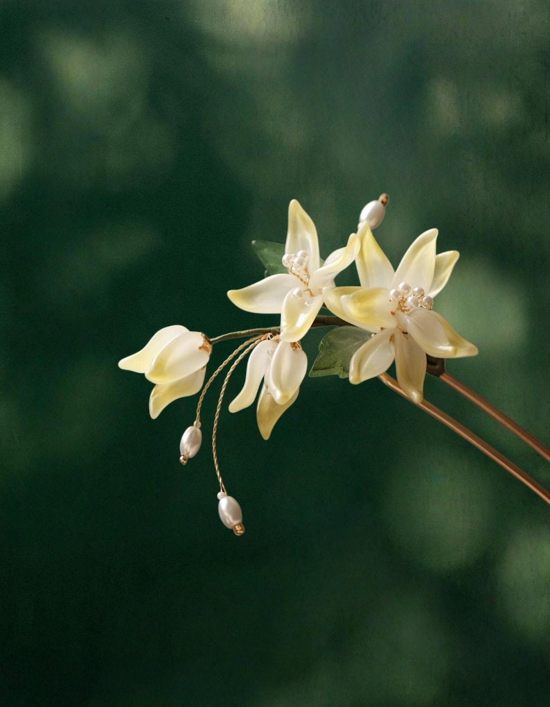 May include: A delicate hair accessory featuring stylized, pale yellow flowers with pearl accents. The floral design includes small green leaves and dangling pearl-like beads, all mounted on a gold-toned metal pin. The background is a blurred, dark green.