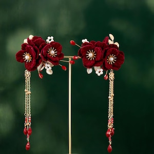 May include: Two ornate hair accessories featuring deep red velvet flowers with gold accents and dangling chains. Each flower has a pearl-like center and is surrounded by gold leaves and small red beads. The chains end with teardrop-shaped red beads.