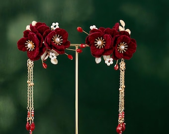 Op de afbeelding: Twee sierlijke haaraccessoires met dieprode fluwelen bloemen met gouden accenten en hangende kettingen. Elke bloem heeft een parelachtig centrum en is omgeven door gouden bladeren en kleine rode kralen. De kettingen eindigen met druppelvormige rode kralen.