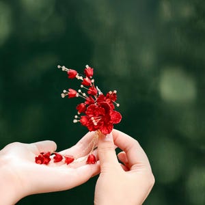 Op de afbeelding: Een levendig rood bloemen haaraccessoire met delicate gouden ketting accenten. Het handgemaakte stuk heeft meerdere kleine bloemknoppen en een grotere centrale bloem. Het accessoire wordt tegen een vage groene achtergrond gehouden.