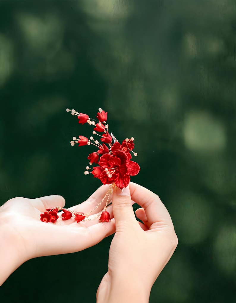 Op de afbeelding: Een levendig rood bloemen haaraccessoire met delicate gouden ketting accenten. Het handgemaakte stuk heeft meerdere kleine bloemknoppen en een grotere centrale bloem. Het accessoire wordt tegen een vage groene achtergrond gehouden.