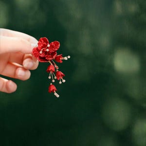 Op de afbeelding: Een rode bloemen haaraccessoire met delicate klokvormige bloemen en kleine witte accenten. Het accessoire wordt tegen een vage groene achtergrond gehouden, wat het ingewikkelde ontwerp en de levendige kleur benadrukt. Geschikt voor speciale gelegenheden.