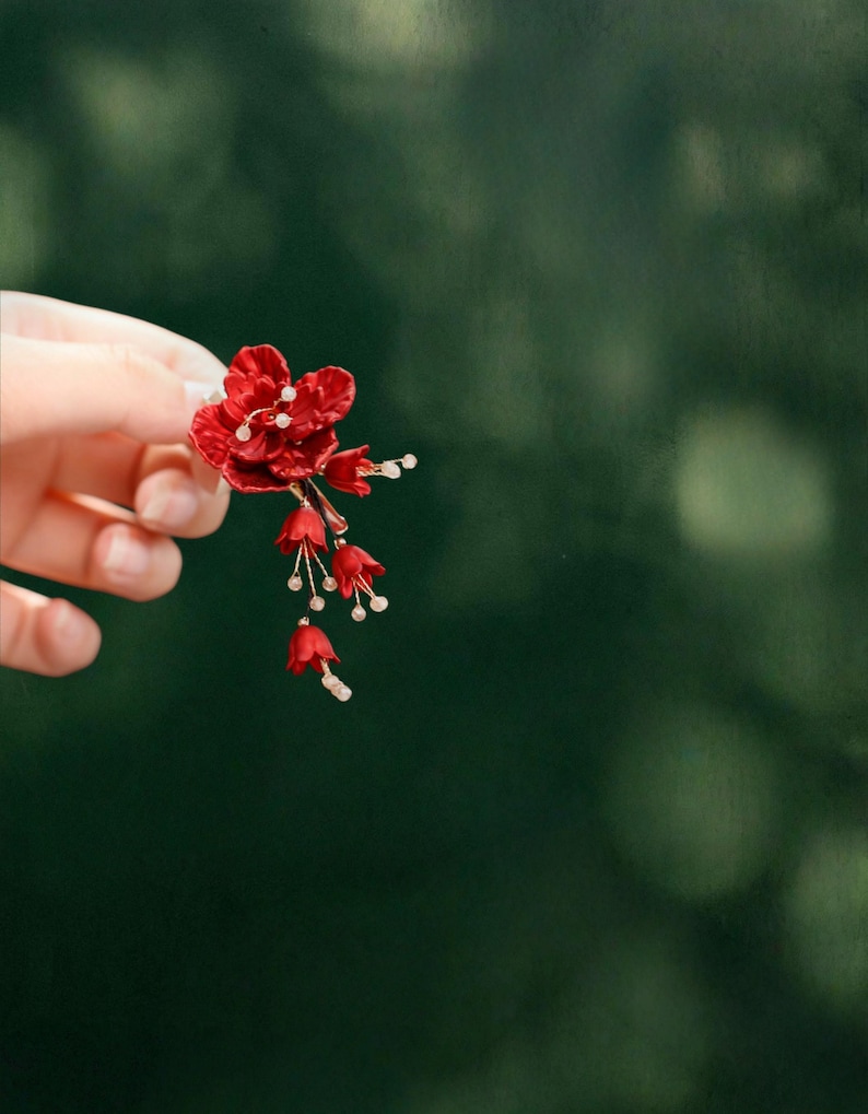 Op de afbeelding: Een rode bloemen haaraccessoire met delicate klokvormige bloemen en kleine witte accenten. Het accessoire wordt tegen een vage groene achtergrond gehouden, wat het ingewikkelde ontwerp en de levendige kleur benadrukt. Geschikt voor speciale gelegenheden.