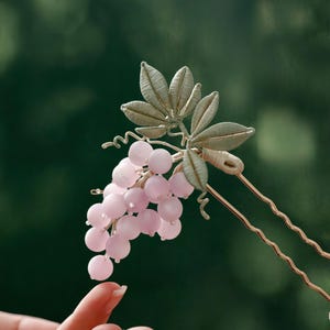 May include: A decorative hair pin featuring a cluster of pink beads resembling grapes, with detailed metallic leaves and vines. The pin has a gold-colored stem and is designed to be inserted into the hair. The overall design is elegant and nature-inspired.