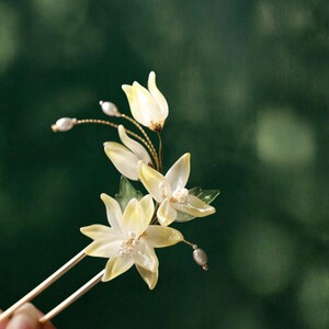 May include: A delicate hair pin featuring translucent, pale yellow flower blossoms with pearl accents. The floral arrangement is mounted on two slender, gold-toned metal prongs, creating an elegant accessory for special occasions.