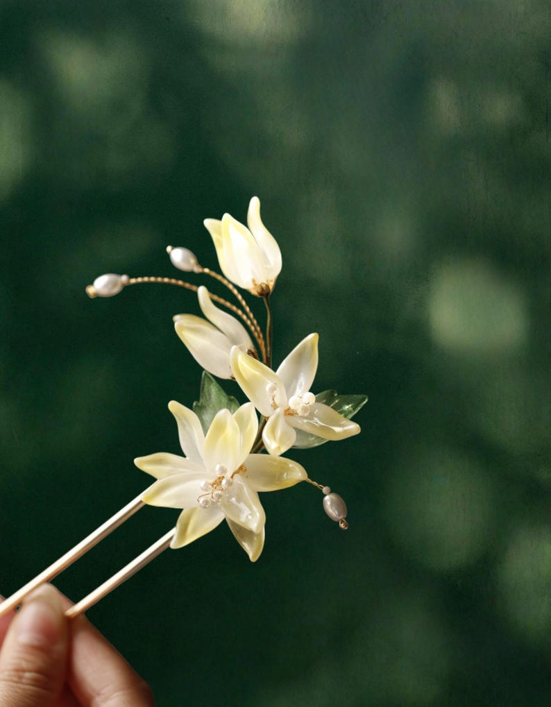 May include: A delicate hair pin featuring translucent, pale yellow flower blossoms with pearl accents. The floral arrangement is mounted on two slender, gold-toned metal prongs, creating an elegant accessory for special occasions.