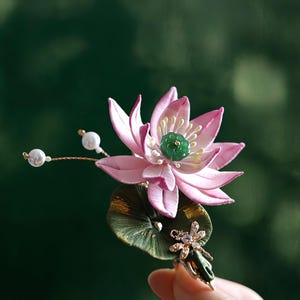 May include: A handcrafted pink lotus flower brooch with a green jade center and a green leaf base. The brooch features delicate pearl accents and a small, jeweled embellishment. The background is a blurred green.