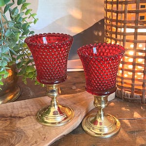 May include: Two red glass candle holders with a textured surface and brass bases. The candle holders are on a wooden surface. The candle holders are a decorative item.
