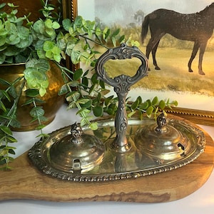 May include: An antique silver-plated serving set with a handle and two lidded compartments. The oval tray sits on a wooden board, with a painting of a horse in the background. Greenery adds a touch of freshness to the scene.