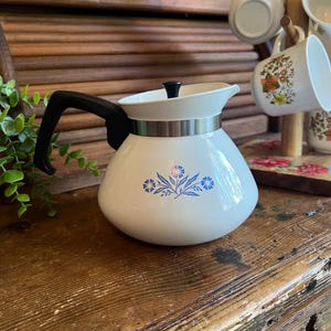 May include: A white teapot with a black handle and a small black lid knob. The body features a blue floral pattern and a silver-colored metal band around the neck. The teapot has a rounded base and a spout integrated into the upper rim design.
