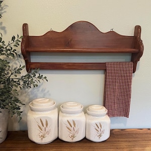 May include: A wooden shelf with a towel rack and three ceramic canisters. The shelf is stained a dark brown and has a decorative top. The canisters are off-white with wheat designs.