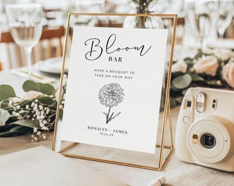 May include: A gold-framed sign with the words "Bloom Bar" and a floral illustration. The sign reads "Make a bouquet to take on your way" and includes names and a date. A white instant camera and a notebook are in the foreground.