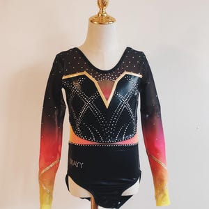 May include: Black gymnastics leotard with long sleeves. The bodice features a geometric design with gold accents and rhinestones. The sleeves transition from black to orange and yellow. The leotard is displayed on a mannequin.