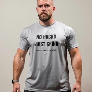 May include: A light gray short-sleeved crew neck t-shirt featuring the black text NO HACKS JUST GRIND printed on the chest, with the smaller text ALPHASET // DEEM READY FIGHT APPAREL printed directly below the main slogan.