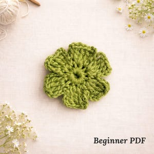May include: A handmade, green crochet flower with five petals. The flower is centered on a cream-colored fabric, with a ball of yarn and a crochet hook in the background. The text "Beginner PDF" is at the bottom.