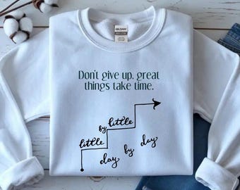 May include: White crewneck sweatshirt with the text "Don't give up, great things take time." in black script. A staircase graphic with the words "little by little, day by day" is also featured. The sweatshirt is folded.