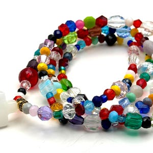 May include: A colorful beaded rosary with a white cross pendant. The beads are a mix of shapes and sizes, including round, faceted, and irregular, in a variety of colors such as red, blue, green, yellow, and purple. The rosary is on a white background.