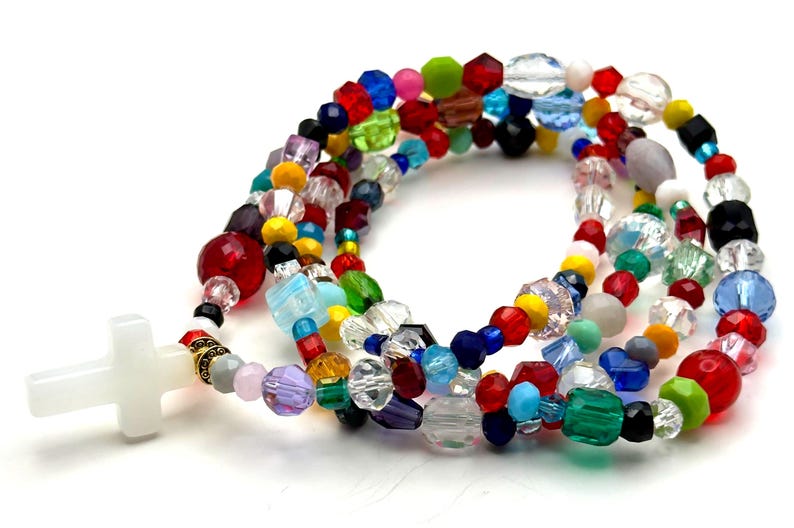 May include: A colorful beaded rosary with a white cross pendant. The beads are a mix of shapes and sizes, including round, faceted, and irregular, in a variety of colors such as red, blue, green, yellow, and purple. The rosary is on a white background.