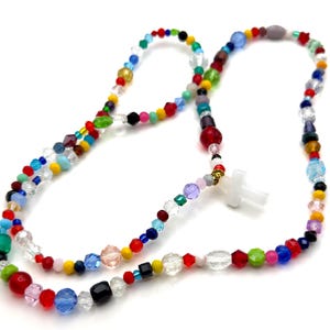 May include: A colorful beaded necklace with a white cross pendant. The necklace features a variety of glass beads in red, blue, green, yellow, and clear. The beads are strung together with a gold-colored metal clasp.