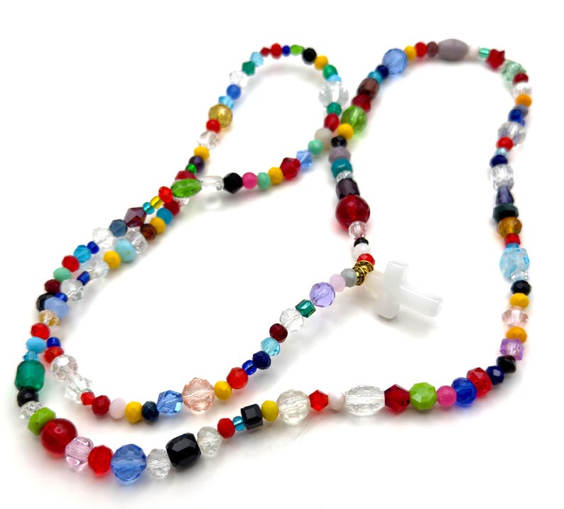 May include: A colorful beaded necklace with a white cross pendant. The necklace features a variety of glass beads in red, blue, green, yellow, and clear. The beads are strung together with a gold-colored metal clasp.