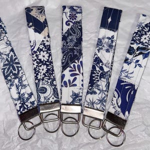 May include: Five fabric wrist keychains with various blue and white floral and paisley patterns. Each keychain has a silver-colored metal ring and a rectangular metal piece. The keychains are arranged on a white, textured surface.