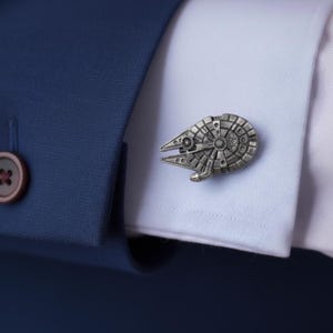 Puede incluir: Gemelo de metal plateado con forma de nave espacial, sujeto a un puño de camisa blanca. El gemelo presenta patrones circulares y rectangulares detallados. Una chaqueta de traje azul marino con un botón marrón en el fondo.