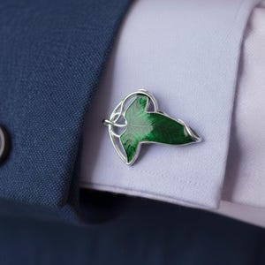 May include: Silver-toned cufflinks featuring a green leaf design. The leaf has a detailed, textured appearance with varying shades of green. The cufflinks are set against a blue suit and a white dress shirt, suggesting formal wear.