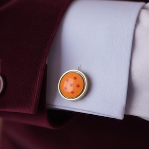 Puede incluir: Gemelos dorados con un centro naranja que presenta cinco estrellas rojas. Los gemelos se colocan contra un puño de camisa azul claro y una manga de chaqueta burdeos, creando un contraste de colores y texturas. Un accesorio elegante.