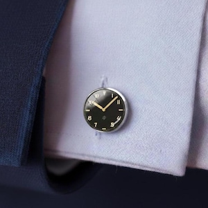 Puede incluir: Gemelos plateados con un diseño de esfera de reloj negra. La esfera del reloj presenta manecillas doradas y números romanos. Los gemelos están sujetos a una camisa azul claro.