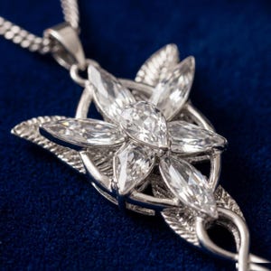 May include: A silver-toned pendant necklace featuring a flower-like design with clear, sparkling gemstones. The pendant has a detailed, leaf-like structure and is suspended from a delicate chain. The necklace is displayed against a dark blue background.
