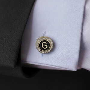 Puede incluir: Gemelo dorado con un centro negro con la letra "G". El gemelo está rodeado por un anillo de pequeños cristales transparentes. El gemelo está sobre una camisa azul claro.