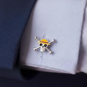 Pode incluir: Abotoaduras prateadas com um design de caveira e ossos cruzados, com um chapéu amarelo e vermelho. As abotoaduras estão em uma manga de camisa azul clara, com um casaco azul marinho ao fundo.
