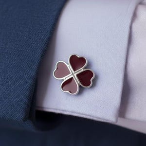 May include: A silver cufflink featuring a four-leaf clover design. The clover's leaves are a deep burgundy, edged in silver. The cufflink is set against a white shirt and a navy blue jacket.