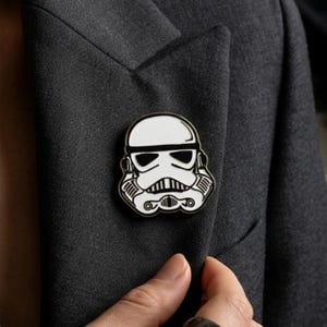 Peut inclure: Épingle en émail noir et blanc représentant un casque de Stormtrooper, fixée sur une veste grise foncée. L'épingle, de forme ronde, présente le design détaillé du casque emblématique de Star Wars. La finition est brillante.