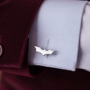 Könnte beinhalten: Silberfarbene Manschettenknöpfe mit dem Fledermaussymbol. Die Manschettenknöpfe sind an einem hellblauen Hemdärmel zu sehen, kombiniert mit einem bordeauxfarbenen Blazer. Das Fledermaussymbol ist ein stilisiertes, symmetrisches Design.