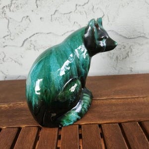 Puede incluir: Figura de zorro de cerámica en tonos verdes y negros. El zorro está sentado con la cabeza girada hacia la derecha. El esmalte tiene un efecto veteado, añadiendo profundidad e interés visual a la escultura. La figura está colocada sobre una superficie de madera.