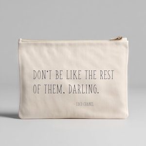 May include: A cream-colored zippered pouch with the quote "DON'T BE LIKE THE REST OF THEM, DARLING." in black text. The quote is attributed to Coco Chanel. The pouch is rectangular and appears to be made of canvas or a similar fabric.