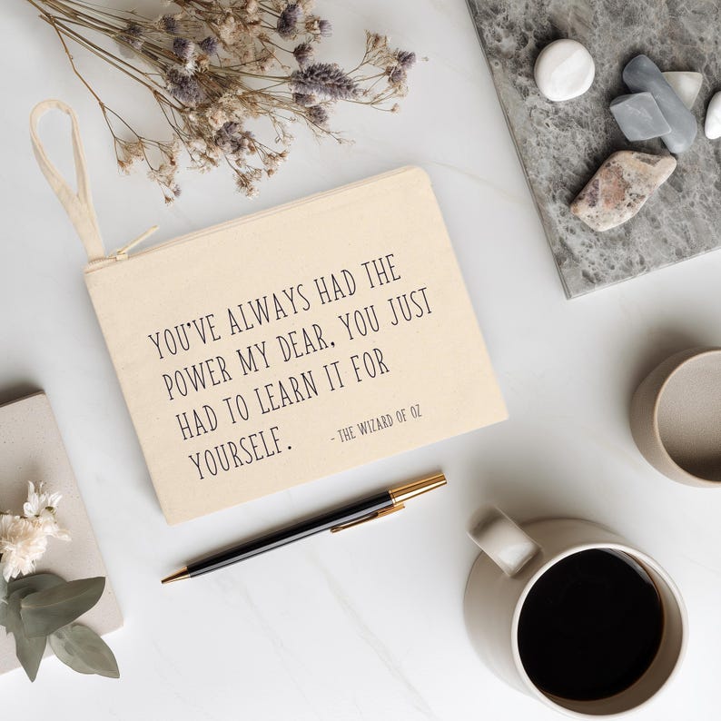 May include: A beige canvas pouch with a zipper and a loop handle. The pouch has the quote "YOU'VE ALWAYS HAD THE POWER MY DEAR. YOU JUST HAD TO LEARN IT FOR YOURSELF." from The Wizard of Oz. A black pen, coffee cup, and dried flowers are also visible.