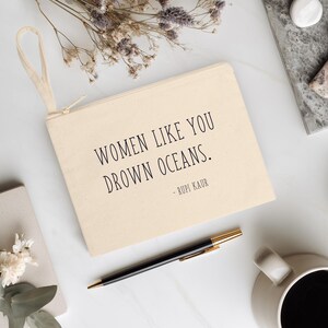 K&ouml;nnte beinhalten: Eine beigefarbene Canvas-Tasche mit Handschlaufe, mit dem Zitat "WOMEN LIKE YOU DROWN OCEANS." von Rupi Kaur in schwarzer Schrift. Die Tasche liegt auf einer wei&szlig;en Oberfl&auml;che mit einem Stift, einer Kaffeetasse und Trockenblumen.