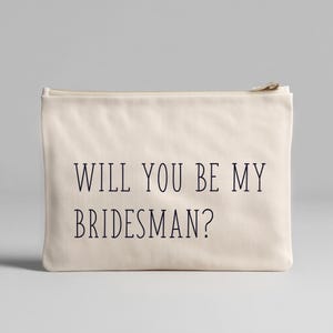 May include: A cream-colored zippered pouch with the text "WILL YOU BE MY BRIDESMAN?" in dark blue. The pouch is rectangular and appears to be made of canvas or a similar fabric. The zipper is a light tan color.