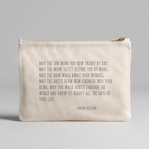 May include: A cream-coloured zippered pouch with the text "MAY THE SUN BRING YOU NEW ENERGY BY DAY...APACHE BLESSING" printed in dark grey. The pouch is rectangular and appears to be made of canvas or a similar fabric.
